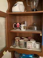 Wood cabinet shelves with a white Osterizer blender base on the top shelf, a clear plastic blender pitcher with lid, and an assortment of coffee mugs on two shelves below. Some mugs have printed logos and designs.