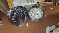 Both 24 inch wall clocks side by side wrapped in plastic, one black face with large white numerals and pendulum, one white face with black numerals and hands.