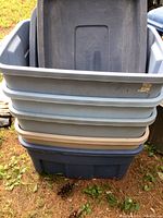 Stack of five Roughneck plastic storage totes of various colors including blue, gray, and tan. One lid missing from bottom tote. Shows outdoor ground background with grass and pinecones.