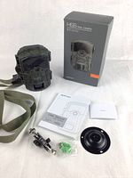 Trail camera in camouflage casing with strap, packaging box, user manual, mounting bracket, wall mount plate, screws, and wall anchors arranged on white surface.