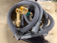 Two drywall sanding heads mounted on wooden bases with yellow plastic handles and green adjustment collars, coiled gray flexible hoses with vacuum connectors nearby on wood surface.