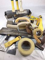 Overview of various drywall finishing and tiling tools including taping knives, drywall tape, and scrapers with visible plaster residue