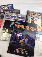 Multiple guitar music books overlapping on a surface, showing titles and partial covers for folk and artist focused collections.