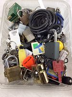 Overhead photo showing a variety of locks and keys in a plastic container. Includes brass and steel padlocks, keychains, and a black coiled cable lock.