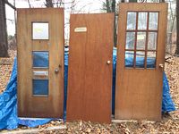 Three doors shown side by side against a blue tarp; one with 3 vertical glass panels and mail slot, one plain wood door, one with 9 glass panes