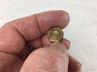 Photo of front side of the gold Paramount Pictures 100% Club lapel pin with diamond set and inscriptions clearly visible held between thumb and forefinger.