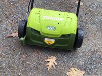 Front view of the SunJoe electric lawn aerator and dethatcher showing the green housing, large wheels, brand logo and safety warning label.
