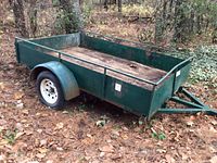 Angled front-left view of green steel utility trailer showing wood deck, side rails, single axle and white wheel; surface rust visible on frame.