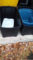 Four assorted plastic storage bins placed on a concrete surface with fallen leaves nearby. Two bins have lids, the small one with a white lid and the larger with a blue lid.