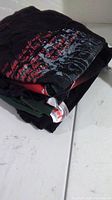 Stack of 5 folded men's graphic t-shirts, showing edges and colors, mostly black/dark with visible red and grey print