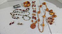 Twelve pieces of costume jewelry arranged on white background. Includes two matching sets with necklaces, bracelets, and earrings in red and turquoise color schemes plus stone bracelets and necklaces with orange and amber tones.