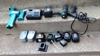 Various cordless tool batteries, battery chargers, and a blue cordless drill frame without battery, arranged on outdoor steps.