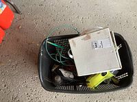 Photo displaying black plastic basket containing the Power Fist 2 ball hitch assembly, booster cables, battery-powered flashlight, and stake with pet leash, with visible rust on hitch component.