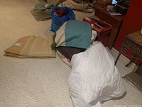 Wide view of assorted bedding items on floor including beige mattress protector, multi-colored throw, white pillow cases, folded bedding items
