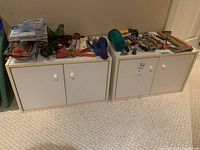 Two white cabinets with doors side-by-side on carpeted floor, with tools displayed on top. Cabinets appear lightly used with smooth finish and white knobs.