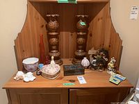 Two photos showing a wooden hutch with an assortment of candle holders, ceramic figurines, decorative boxes, glass bottles, and magnets arranged on its shelves.