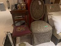 Two different chairs side by side; one upholstered in patterned fabric with a rounded back and skirted bottom, the other wooden framed with a floral needlepoint seat.