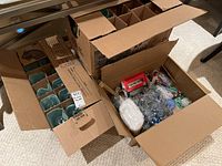 Four open cardboard boxes containing Christmas glassware and holiday decorations. One box contains blue glassware items individually packed; another box contains various decorations including tinsel, ornaments, and a small red nativity set.