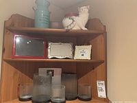 Top wooden shelf displaying three photo frames, five ribbed glass candle holders, and white ceramic cat candle holder next to blue tin vase.