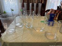 Five large clear glass vases of mixed cylindrical and hourglass shapes arranged on a table with a light green tablecloth.