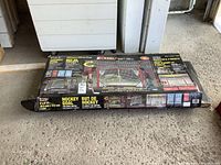 Box containing folding metal hockey goal with backstop and four targets, shown on concrete floor, unopened original packaging.