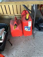 Two red resin jerry cans side by side under a workbench on a concrete floor, showing yellow spouts and handles.