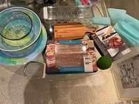 Overview of kitchenware items including nested bowls, wooden cutting board, pink salt plate in packaging, silicone food storage bags, egg crate, and poultry lifter tool