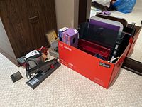 Photo showing a variety of office supplies including staplers, a red box filled with folders, and packs of paper clips and bulldog clips on carpeted floor.
