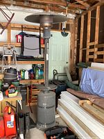 Full view of the propane patio heater standing upright in a garage, showing the base with wheels, control panel, tall heating element tube, and round reflector on top.