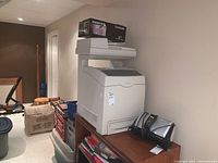 Front view of the Lexmark X560n multifunction printer on a wooden cabinet, with a printer cartridge box and other items placed on top, surrounded by boxes and household items.