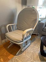 Front angled view of white rocking glider chair with patterned beige cushions on wooden floor next to a rug and grey sofa.