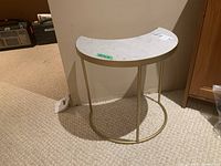 Side view of half-moon side table with white stone top and gold-tone metal base against beige carpet and wall.