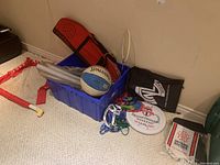 Room corner with blue storage bin containing badminton rackets, paddles, a white and blue Spalding basketball needing air, pennants, colorful lanyards, and assorted cords. Red and white badminton net and poles on the carpet floor nearby.