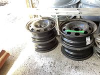 Four black steel rims stacked two on two on concrete floor in garage area. Rust and surface wear visible. Light source from open garage door.