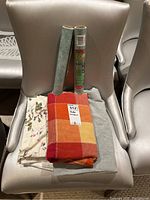 Photo showing three folded table linens on a chair: a patterned linen with autumn leaves, a checkered red and orange linen, and a gray linen, with two standing rolls of Contact brand paper liner.