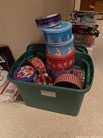 Green plastic storage tub filled with 15 assorted decorative tins of various shapes and sizes. Common holiday and winter-themed designs visible including snowmen, Christmas trees, and ornaments.