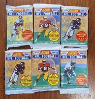 Six unopened 1990 Score Series 2 NFL Football trading card packs displayed on a wooden surface, each featuring an NFL player image and promotional stickers.