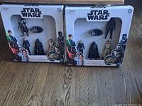 Two unopened boxes of Star Wars figures, each box contains four visible action figures and a small accessory, side-by-side on wooden floor.