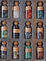 Top view of 12 small glass bottles with cork stoppers, each filled with different polished gemstones and labeled: Green Agate, Opal, Red Coral, Blue Apatite, Cat's Eye, Obsidian, Sodalite, Strawberry Quartz, Lapis Lazuli, White Agate, Green Turquoise, Goldstone.
