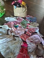 Photo showing assortment of vintage doll clothes spread out next to a beige bag filled with more doll clothes and accessories.
