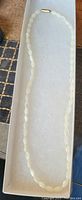 Photo shows a single strand necklace made of oval mother of pearl beads laid out in a white box liner, approximately 16 inches long, without box lid.