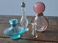 Front and side view of four vintage perfume bottles including two clear textured glass bottles with silver pointed stoppers, one turquoise blue glass bottle with rounded stopper, and one round pink glass bottle with large round pink stopper on rose gold collar.
