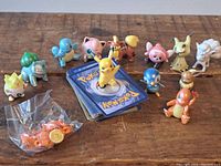 Photo showing 13 small Pokemon figurines including Pikachu, Charmander, Bulbasaur, Squirtle, and others, alongside a stack of Pokemon cards and a small bag with an orange item.