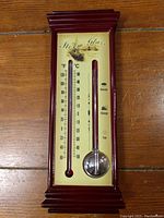 Front view of the storm glass and thermometer unit showing the wood frame and yellow background panel with thermometer and storm glass tube