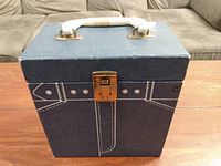 Front view of blue fabric-covered cube-shaped vintage travel record storage box with white handle and brass latch, showing some wear.