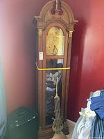 Front view of the tall wooden Pearl grandfather clock standing in a corner with visible brass accented dial and glass door, partially disassembled with pendulum held by rubber band.