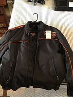 Black Harley Davidson jacket laid flat showing front view with zipper closed, orange piping details on sleeves and chest logo.