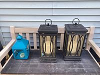 Two black battery-operated lanterns and one blue metal candle lantern on wooden bench.