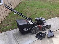 Full view of Craftsman lawnmower with grass collection bag, handlebar, and extra attachments placed beside it on pavement and grass background.