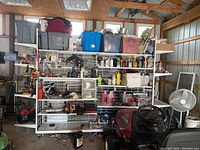 Wide view of the white metal shelving unit loaded with miscellaneous items stored on shelves and hooks, set inside a garage or workshop area.